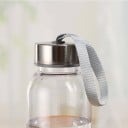 Bouteille de voyage en plastique transparent avec un couvercle à vis argenté et une anse en textile. Forme cylindrique élancée. Pour l'école 4